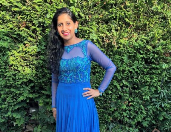 Minreet standing in front of a large green bush, wearing a long blue dress