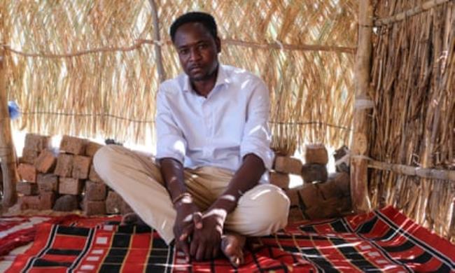 Sherif al-Deen, a social worker, sits in a grass hut on a red woven mat