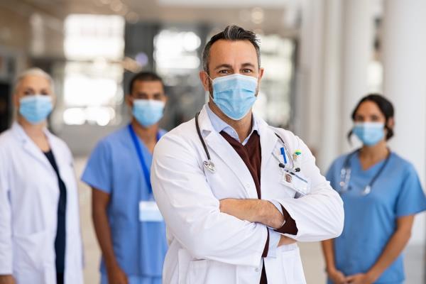 Mature doctor standing in corridor with medical team at hospital wearing surgical face mask due to covid. Smiling general practitio<em></em>ner with crossed arms looking at camera.