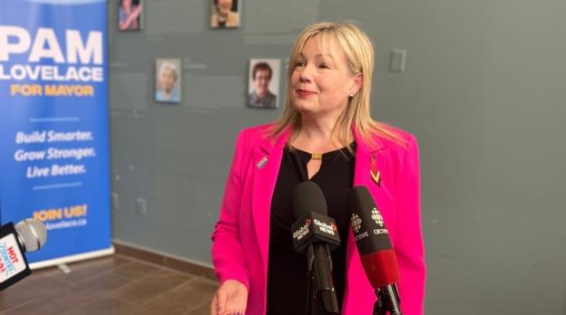 A woman in a black dress and a bright pink blazer stands in front of two micropho<em></em>nes in a grey room.