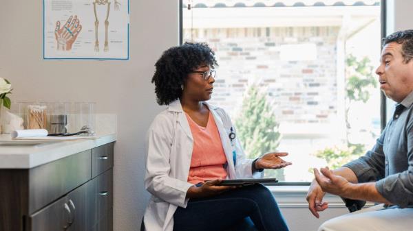 A female physician in a lab coat and pink top talks to a male patient.