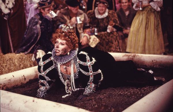 Joan Collins wearing a wig and crown, in a mud pit