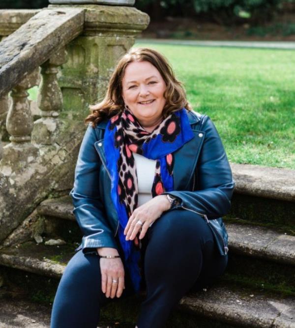 Alison Rios McCrone sitting on stone steps outside, with a stone balustrade to her right and grass in the background.