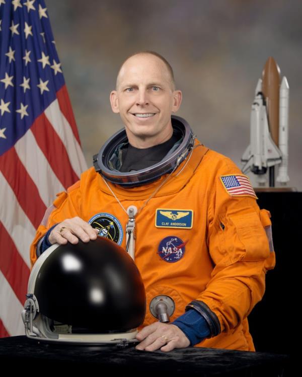 Clayton Anderson, in NASA astro<em></em>naut uniform, posing with his helmet in front of an American flag and spaceship model.