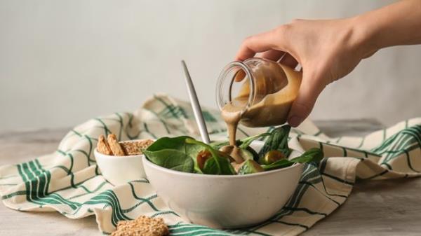 Pouring of tasty tahini from jar o<em></em>nto fresh vegetables in bowl