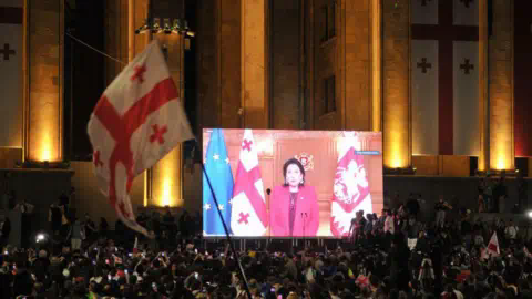 Getty Images A large crowd gathers at night in front of the Georgian Parliament building in Tbilisi, with the illuminated facade adorned with Georgian flags. A giant screen displays President Salome Zourabichvili delivering a speech, standing between the Georgian and European Unio<em></em>n flags.